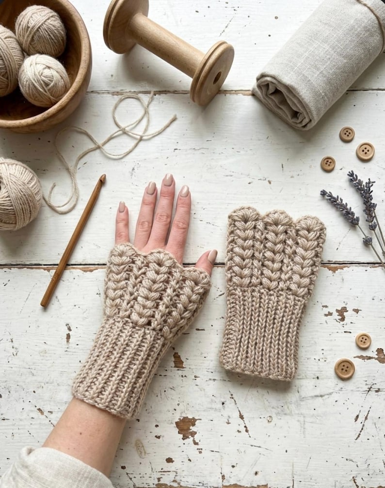 May include: A pair of beige fingerless gloves with a textured, woven design. The gloves are displayed on a white wooden surface, alongside yarn, a crochet hook, wooden buttons, and lavender sprigs. The gloves are a cozy accessory.
