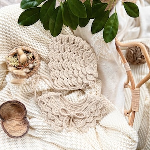 May include: A set of two crocheted baby accessories in a cream color. The set includes a bonnet and a collar with a bow. The accessories are laid out on a white knitted blanket.