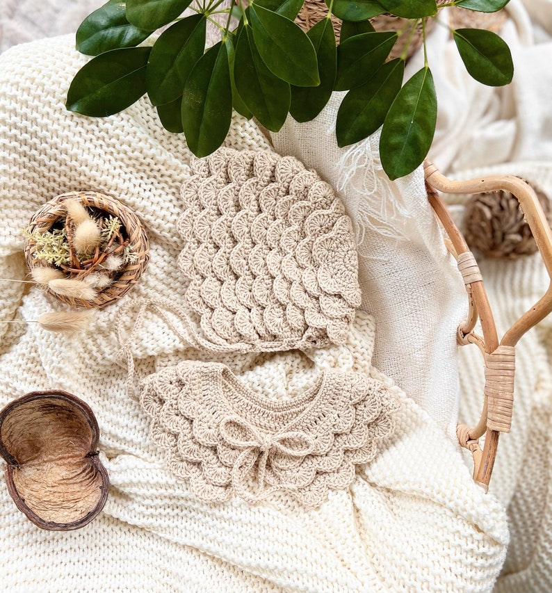 May include: A set of two crocheted baby accessories in a cream color. The set includes a bonnet and a collar with a bow. The accessories are laid out on a white knitted blanket.