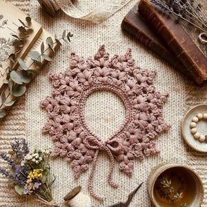 Op de afbeelding: Een stoffige roze gehaakte kraag met een bloemenpatroon en een striksluiting. De kraag is omgeven door vintage items, waaronder boeken, gedroogde bloemen en een kopje thee, alles op een crèmekleurig gebreid oppervlak.