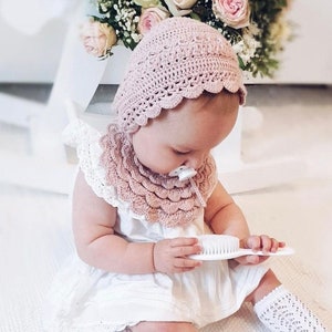 Puede incluir: Un beb&eacute; con un gorro y babero de ganchillo rosa claro, un vestido blanco y calcetines hasta la rodilla. El beb&eacute; sostiene un cepillo blanco. Un ramo de rosas rosas y blancas est&aacute; en el fondo.
