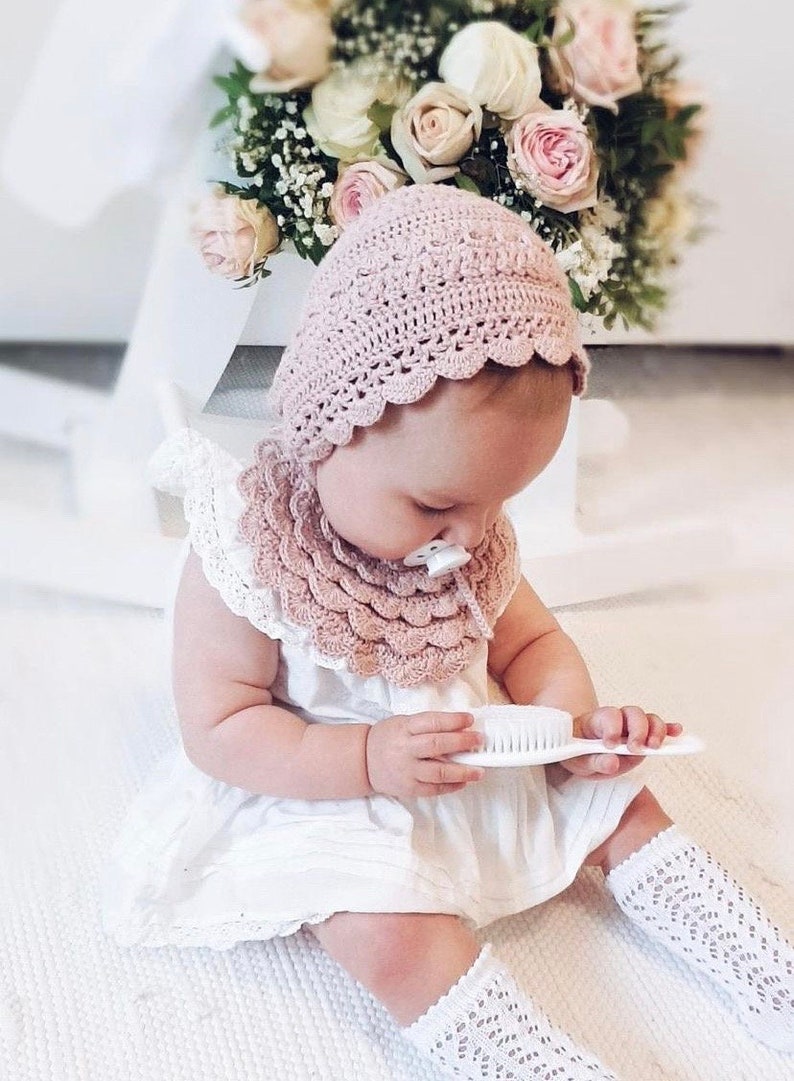 Puede incluir: Un beb&eacute; con un gorro y babero de ganchillo rosa claro, un vestido blanco y calcetines hasta la rodilla. El beb&eacute; sostiene un cepillo blanco. Un ramo de rosas rosas y blancas est&aacute; en el fondo.