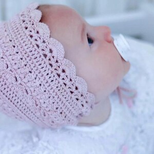 May include: A baby wearing a light pink, crocheted bonnet with a scalloped edge. The baby is also wearing a white dress and has a pacifier in their mouth. The bonnet is made of a soft, textured yarn.