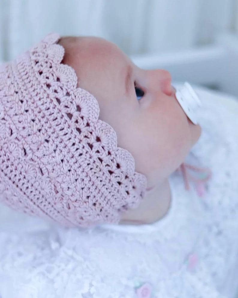 May include: A baby wearing a light pink, crocheted bonnet with a scalloped edge. The baby is also wearing a white dress and has a pacifier in their mouth. The bonnet is made of a soft, textured yarn.