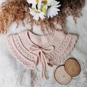 May include: A light pink, crocheted collar with a scalloped edge and a delicate bow tie closure. The collar is displayed on a white, textured surface, with dried wheat stalks and white flowers in the background. Two small wooden slices are also present.