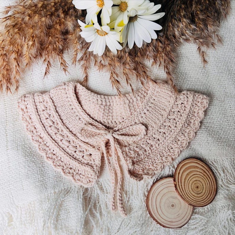May include: A light pink, crocheted collar with a scalloped edge and a delicate bow tie closure. The collar is displayed on a white, textured surface, with dried wheat stalks and white flowers in the background. Two small wooden slices are also present.