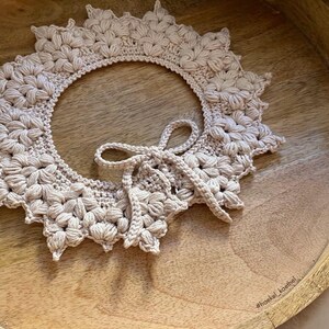 May include: A beige, handmade crochet collar with a floral design and a bow closure. The collar is circular with a scalloped edge and is displayed on a wooden tray. The hashtag #haekel_koethel is visible.