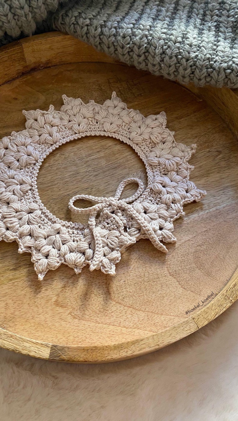 May include: A beige, handmade crochet collar with a floral design and a bow closure. The collar is circular with a scalloped edge and is displayed on a wooden tray. The hashtag #haekel_koethel is visible.