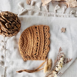 Puede incluir: Un gorro de ganchillo marr&oacute;n c&aacute;lido, con un borde festoneado y delicados detalles calados. El gorro, con lazos, se muestra sobre una superficie texturizada blanquecina. Flores secas y una hoja de madera adornan la escena.