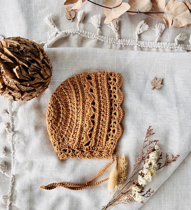 Puede incluir: Un gorro de ganchillo marr&oacute;n c&aacute;lido, con un borde festoneado y delicados detalles calados. El gorro, con lazos, se muestra sobre una superficie texturizada blanquecina. Flores secas y una hoja de madera adornan la escena.