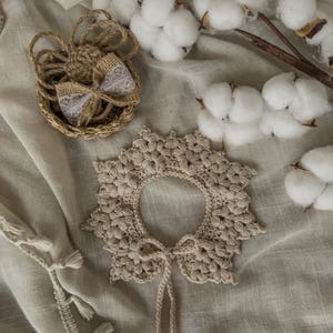 May include: A beige, crocheted collar with a floral design and a matching tie. A small woven basket with a burlap bow and cotton branches are also visible. The items are arranged on a neutral-colored fabric.