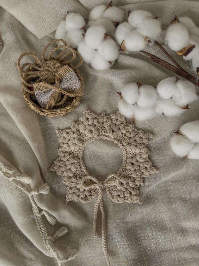 May include: A beige, crocheted collar with a floral design and a matching tie. A small woven basket with a burlap bow and cotton branches are also visible. The items are arranged on a neutral-colored fabric.