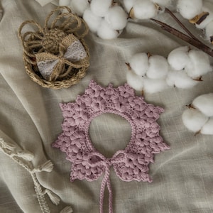 May include: A handmade, light purple crochet collar with a scalloped edge and a tie closure. The collar is surrounded by cotton bolls and a small woven basket with a lace bow. The items are arranged on a neutral-toned fabric.