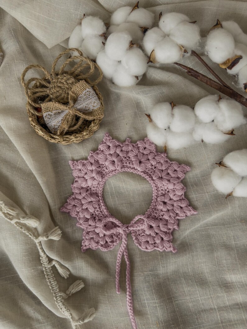 May include: A handmade, light purple crochet collar with a scalloped edge and a tie closure. The collar is surrounded by cotton bolls and a small woven basket with a lace bow. The items are arranged on a neutral-toned fabric.