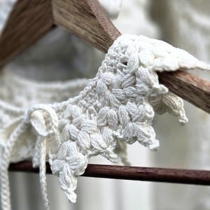 Può includere: Primo piano di un colletto all'uncinetto bianco fatto a mano con un motivo floreale. Il colletto è esposto su una gruccia di legno, mostrando gli intricati dettagli del filato bianco sporco strutturato. Il design include delicati lacci.