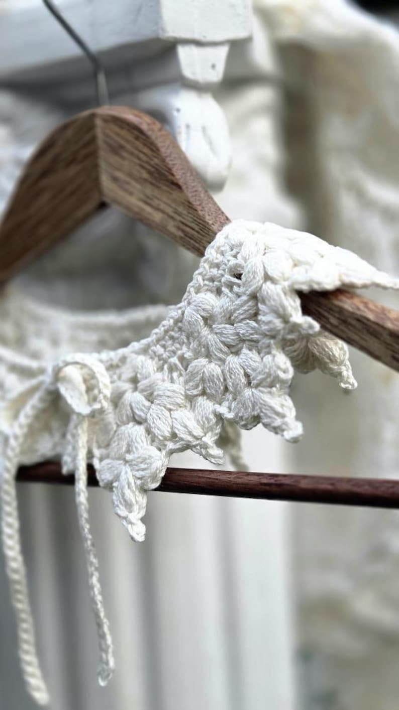 May include: Close-up of a white, handmade crochet collar with a floral pattern. The collar is displayed on a wooden hanger, showcasing the intricate details of the textured, off-white yarn. The design includes delicate ties.