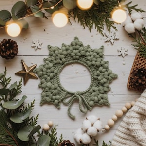May include: A sage green crocheted wreath with a circular center, tied with a bow. The wreath is surrounded by Christmas decorations, including pinecones, a gold star, and sprigs of greenery. Fairy lights and cotton bolls add to the festive scene.