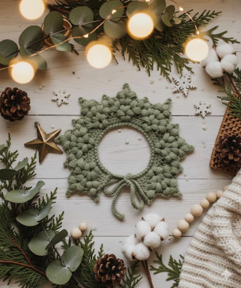 May include: A sage green crocheted wreath with a circular center, tied with a bow. The wreath is surrounded by Christmas decorations, including pinecones, a gold star, and sprigs of greenery. Fairy lights and cotton bolls add to the festive scene.