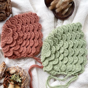 May include: Two crocheted baby hats, one in a light green color and one in a terracotta color. Both hats have a unique scale-like pattern.