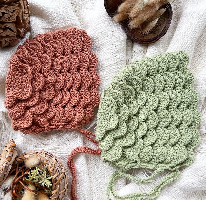 May include: Two crocheted baby hats, one in a light green color and one in a terracotta color. Both hats have a unique scale-like pattern.
