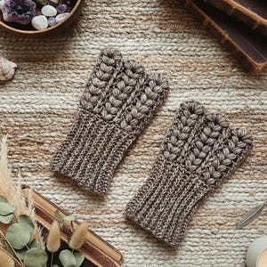 May include: Two brown fingerless gloves with a textured, knitted design. The gloves have a ribbed cuff and a detailed pattern on the top. The gloves are displayed on a textured, neutral-toned surface, suggesting a cozy, handmade aesthetic.