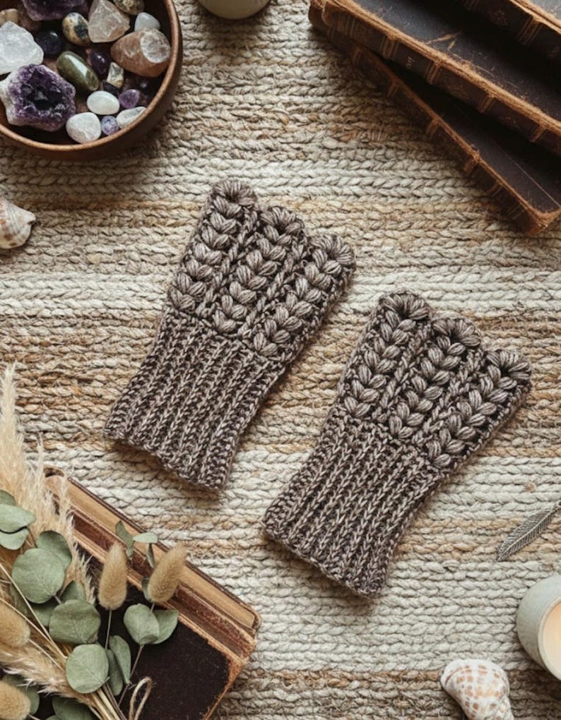 May include: Two brown fingerless gloves with a textured, knitted design. The gloves have a ribbed cuff and a detailed pattern on the top. The gloves are displayed on a textured, neutral-toned surface, suggesting a cozy, handmade aesthetic.