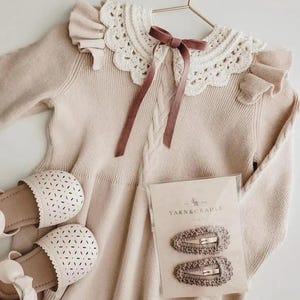 May include: A beige knit dress with a white lace collar and a mauve ribbon. The dress is paired with matching sandals and two hair clips from Yarn & Cradle. The dress is hanging on a gold hanger.
