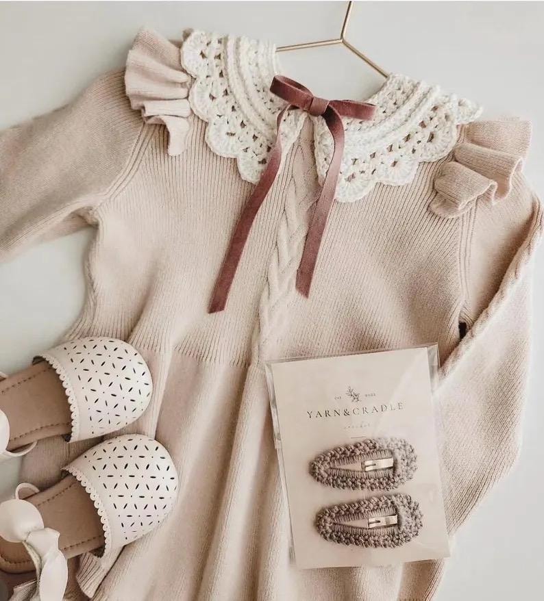 May include: A beige knit dress with a white lace collar and a mauve ribbon. The dress is paired with matching sandals and two hair clips from Yarn & Cradle. The dress is hanging on a gold hanger.