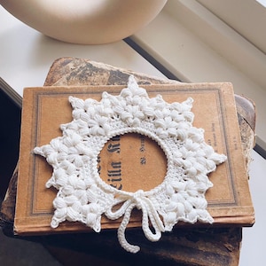 May include: A white, crocheted collar with a floral design and a tie closure. The collar is placed on top of an antique book with faded text. A curved, off-white decorative object is in the background.