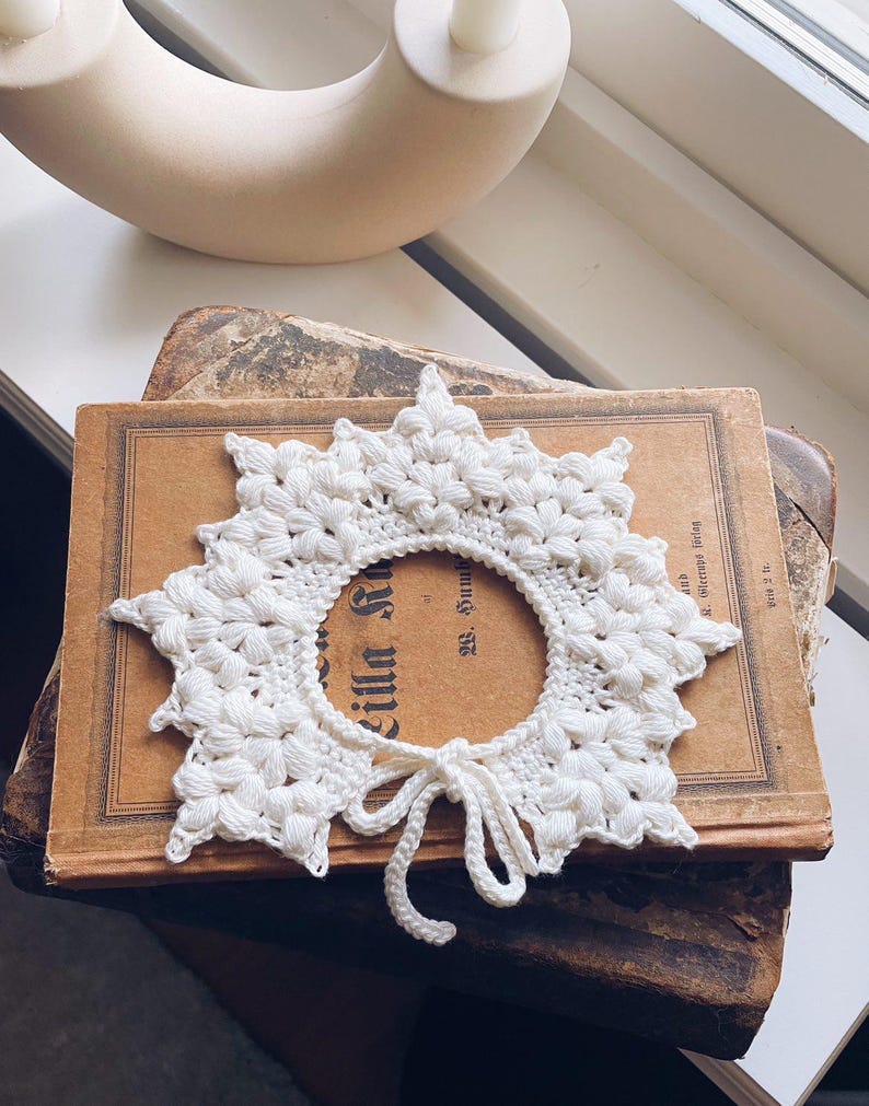 May include: A white, crocheted collar with a floral design and a tie closure. The collar is placed on top of an antique book with faded text. A curved, off-white decorative object is in the background.