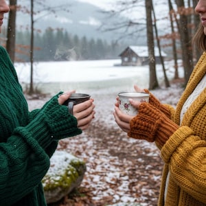 May include: Two people holding mugs of hot beverages in a snowy forest. One person wears a green sweater and the other a yellow cardigan with orange fingerless gloves. The mugs have steam rising from them.