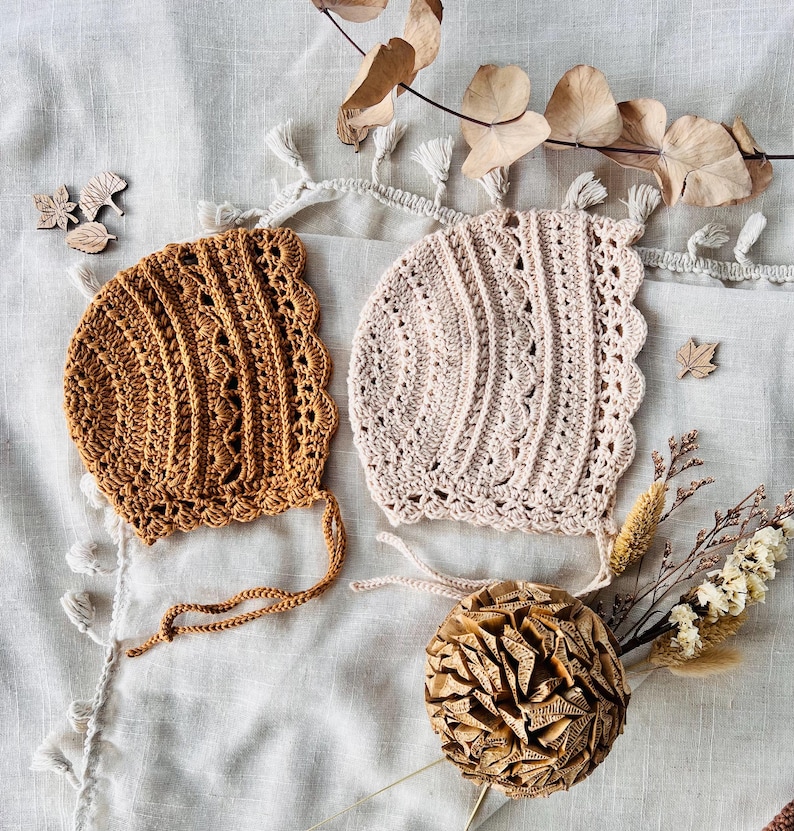 Puede incluir: Dos gorros de beb&eacute; hechos a mano con ganchillo, en tonos beige y marr&oacute;n claro. Los gorros presentan un borde festoneado y delicados patrones calados. La imagen est&aacute; decorada con hojas secas y recortes de hojas de madera, creando una est&eacute;tica r&uacute;stica y oto&ntilde;al.