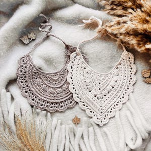 May include: Two handmade crochet bibs in neutral tones. One is a light beige, the other a taupe color. Both have a scalloped edge and a tie closure. The bibs are displayed on a soft, textured surface with decorative elements.