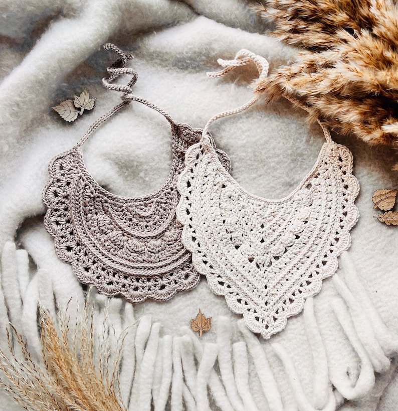 May include: Two handmade crochet bibs in neutral tones. One is a light beige, the other a taupe color. Both have a scalloped edge and a tie closure. The bibs are displayed on a soft, textured surface with decorative elements.
