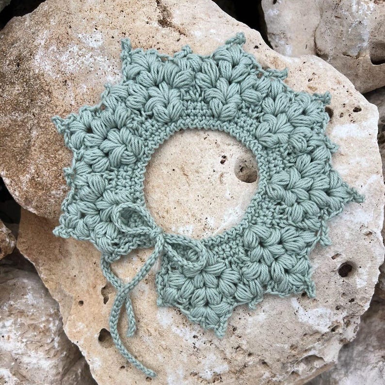 May include: A sage green, handmade crochet collar with a floral pattern. The collar has a round neck opening and a tie closure. The collar is made of soft yarn and is displayed on a textured stone surface.
