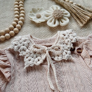 May include: A light pink baby dress with a crocheted collar and a bow. The dress has a textured pattern and small wooden buttons. Two crocheted flower hair clips and a wooden bead garland are also visible.