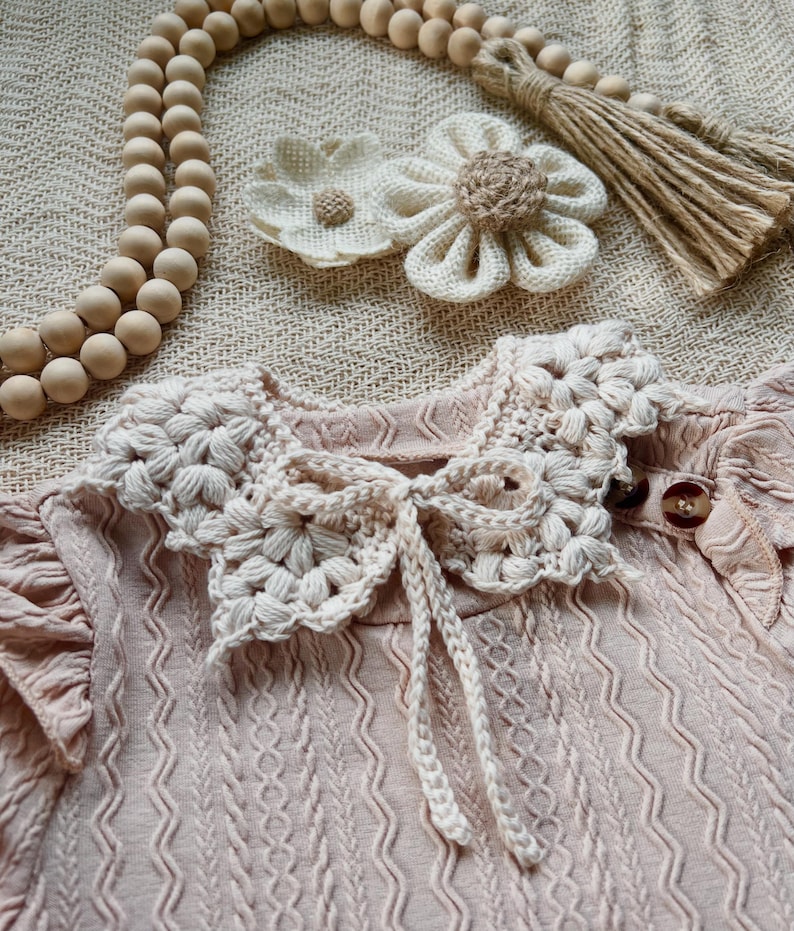 May include: A light pink baby dress with a crocheted collar and a bow. The dress has a textured pattern and small wooden buttons. Two crocheted flower hair clips and a wooden bead garland are also visible.