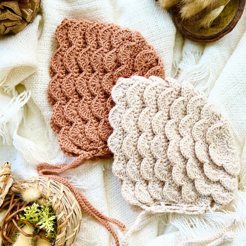 May include: Two crocheted baby bonnets in a beige and terracotta color scheme. The bonnets are made with a textured, scalloped stitch.
