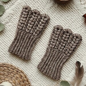 May include: A pair of brown fingerless gloves with a textured, braided design. The gloves are made of a soft, knitted material and are displayed on a cream-colored, textured surface. The gloves are designed to keep hands warm while leaving fingers free.