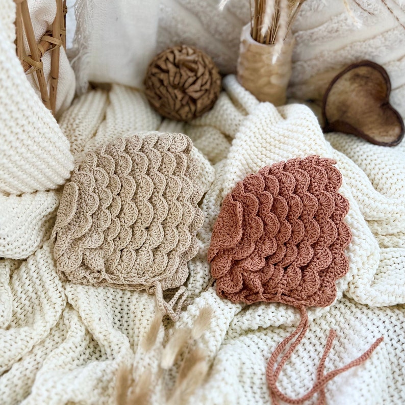 May include: Two crocheted baby bonnets, one in a light brown color and the other in a terracotta color, with a scalloped edge design. The bonnets are lying on a white knitted blanket.