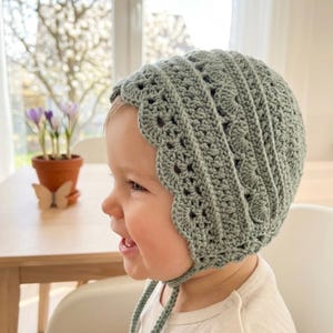 Puede incluir: Un gorro de ganchillo verde salvia con un borde festoneado y lazos. El gorro est&aacute; hecho a mano y parece ser para un beb&eacute; o ni&ntilde;o peque&ntilde;o. El punto de ganchillo crea un dise&ntilde;o abierto y aireado.