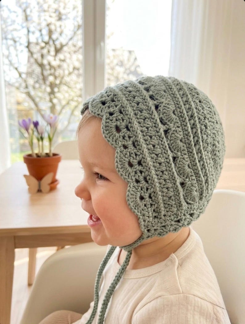 Puede incluir: Un gorro de ganchillo verde salvia con un borde festoneado y lazos. El gorro est&aacute; hecho a mano y parece ser para un beb&eacute; o ni&ntilde;o peque&ntilde;o. El punto de ganchillo crea un dise&ntilde;o abierto y aireado.