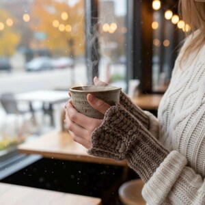 May include: A warm, cozy scene featuring a steaming ceramic mug held by a person wearing a cream-colored cable-knit sweater and brown knitted arm warmers. The background shows a window with a blurred outdoor view, suggesting a cafe setting.