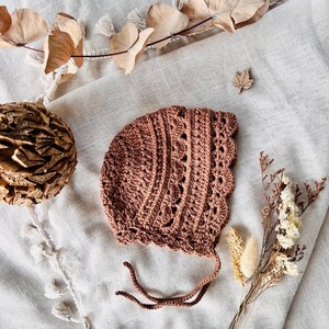 Puede incluir: Un gorro de beb&eacute; de ganchillo en color rosa apagado. El gorro presenta un borde festoneado y patrones de puntadas intrincados. Tiene lazos delicados y se exhibe sobre una superficie de lino neutra, con acentos florales secos.