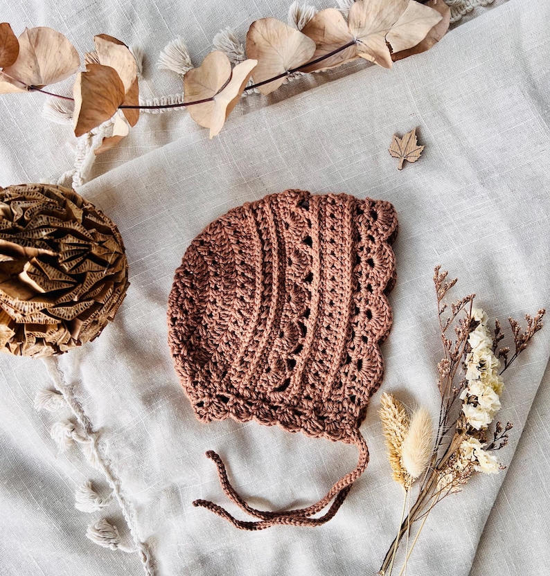 Puede incluir: Un gorro de beb&eacute; de ganchillo en color rosa apagado. El gorro presenta un borde festoneado y patrones de puntadas intrincados. Tiene lazos delicados y se exhibe sobre una superficie de lino neutra, con acentos florales secos.