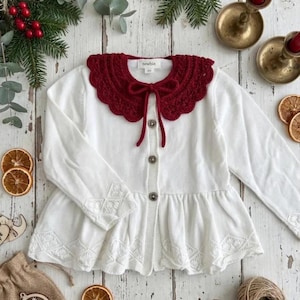 May include: A white baby cardigan with a scalloped, burgundy crochet collar and matching tie. The long-sleeved sweater has decorative embroidery on the cuffs and hem. The cardigan has three buttons down the front.