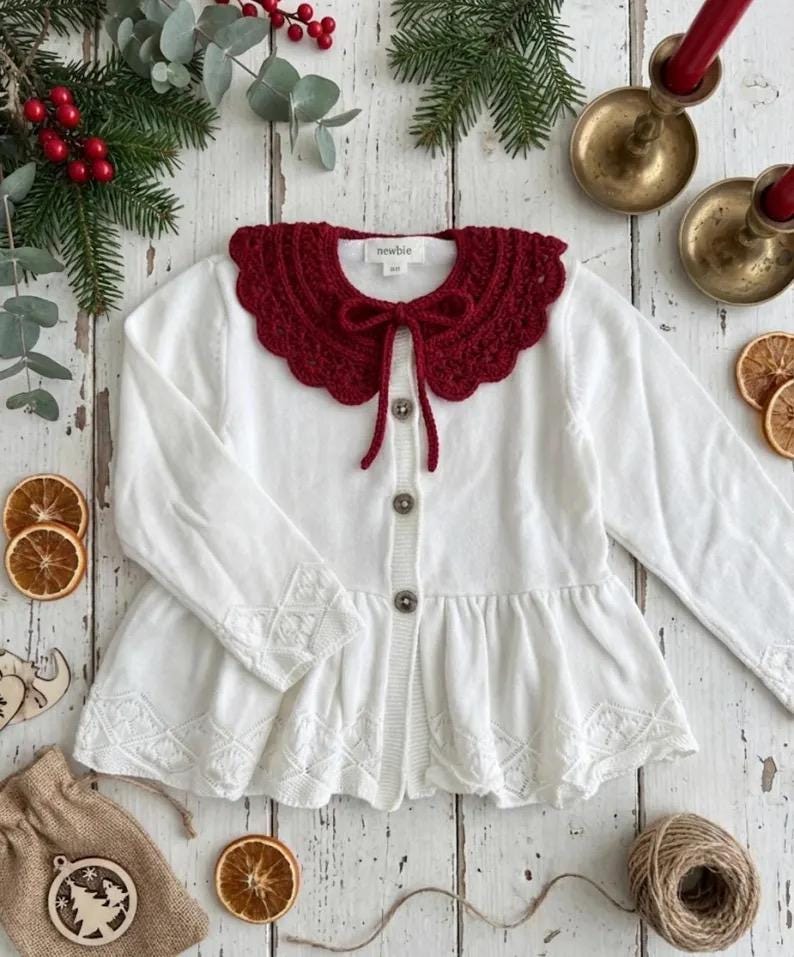 May include: A white baby cardigan with a scalloped, burgundy crochet collar and matching tie. The long-sleeved sweater has decorative embroidery on the cuffs and hem. The cardigan has three buttons down the front.