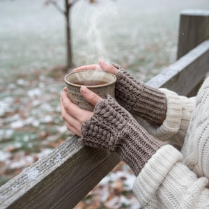 May include: A person holding a steaming ceramic mug, wearing brown knitted fingerless gloves and a cream-colored sweater. The mug is filled with a dark liquid, and the scene is set outdoors with a snowy backdrop and a wooden fence.