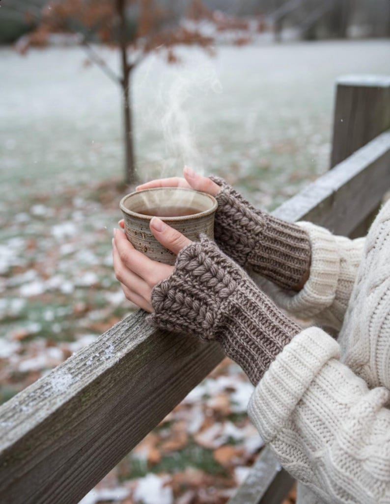 May include: A person holding a steaming ceramic mug, wearing brown knitted fingerless gloves and a cream-colored sweater. The mug is filled with a dark liquid, and the scene is set outdoors with a snowy backdrop and a wooden fence.