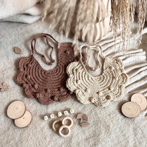 May include: Two crocheted bibs with a scalloped edge and a textured design. The bibs are in brown and beige and have a daisy charm.